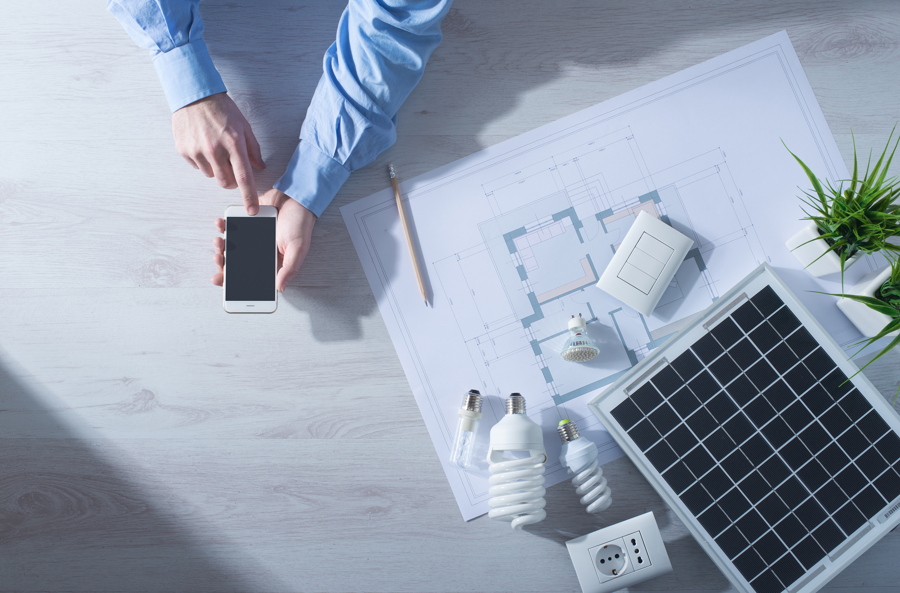 Hand holding smart phone on top of desk with new energy efficient light bulbs and blue prints